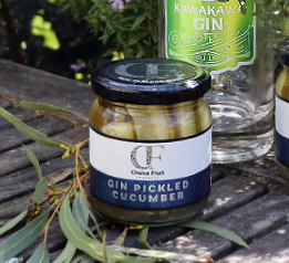 A jar of Gin Pickled Cucumber placed on a wooden surface, with a bottle of gin and some foliage in the background.