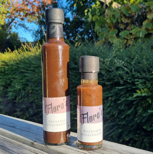Two bottles of Flora's Balsamic Vinaigrette, one larger and one smaller, placed on a wooden surface with foliage in the background.