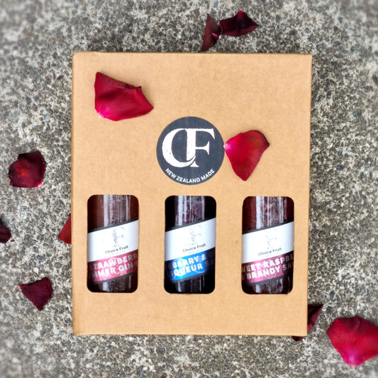 Three small bottles of gin in a cardboard box with rose petals on a stone surface.