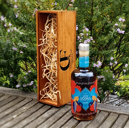 A bottle of Koakoa Coffee Vodka with a colorful label, alongside two clear pressed glass tumblers, all presented in a stained wooden gift box with a logo on the front.