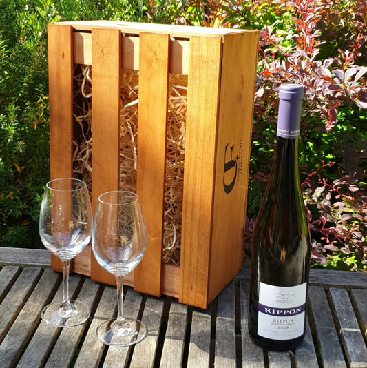 Wine bottle with a purple label, two wine glasses, and a wooden crate on a wooden table outdoors.