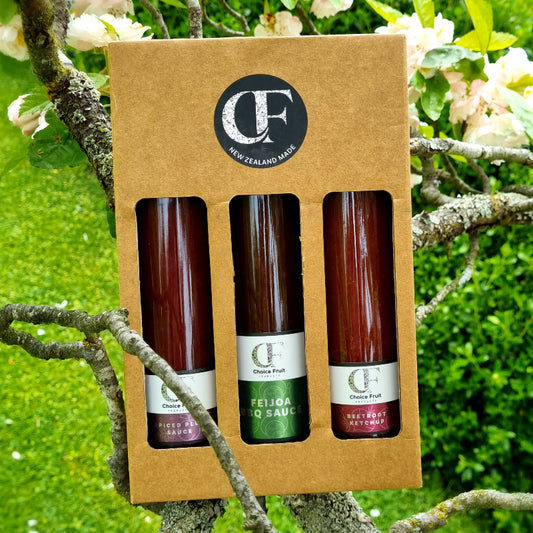 Three bottles of sauce in a cardboard box with a brand logo, set against a natural background.