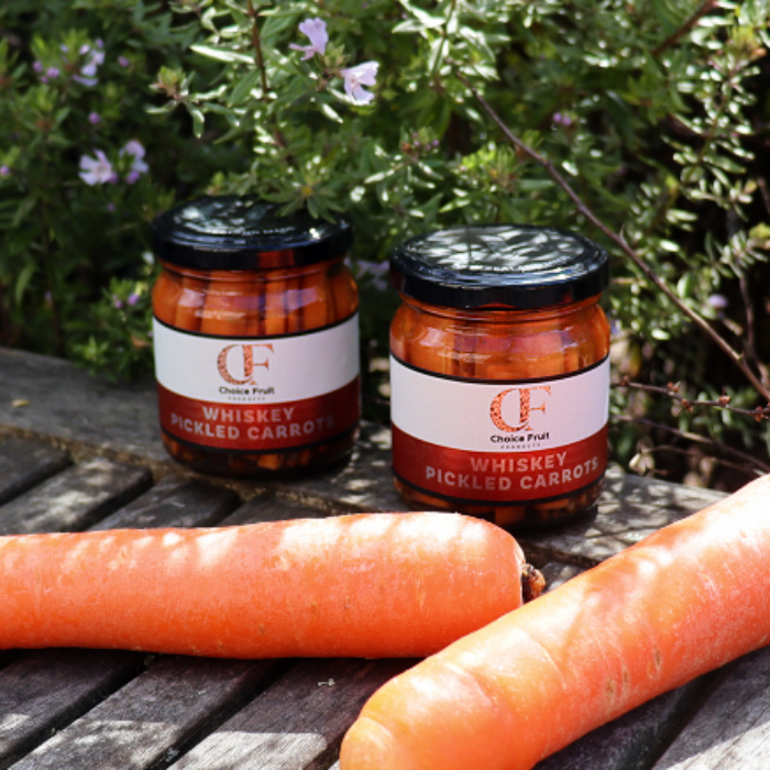 Two jars of whiskey pickled carrots on a wooden surface with carrots in front, against a natural background.