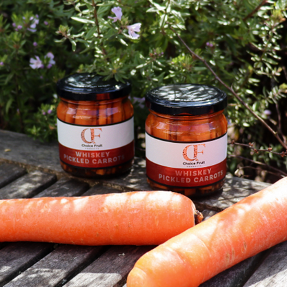 Two jars of whiskey pickled carrots on a wooden surface with carrots in front, against a natural background.