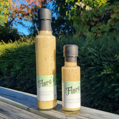 Two bottles of Flora's Apple Cider vinaigrette dressing, one larger and one smaller, with a blurred background featuring greenery and a blue sky.