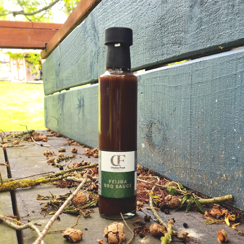 Bottle of Feijoa BBQ Sauce placed upright on a wooden surface with autumn leaves scattered around, under a blue painted wooden cart.