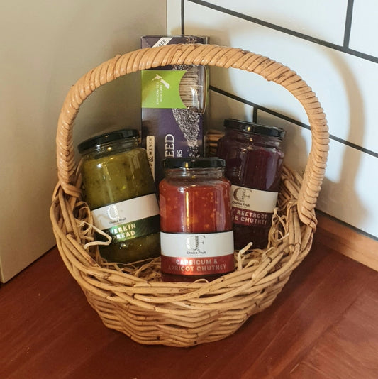 A wicker gift basket containing jars of chutney and a box of crackers, all decorated with a ribbon bow.