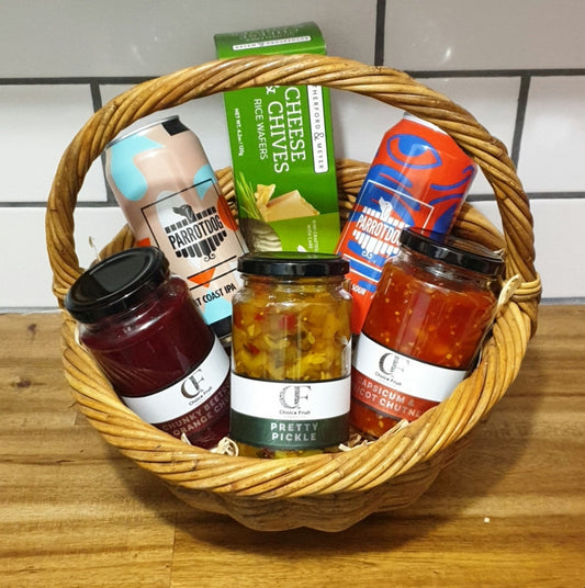 A wicker gift basket containing a variety of food items including jars of beetroot, orange, and apricot products, a can of Parrotdog beer, and rice crackers.