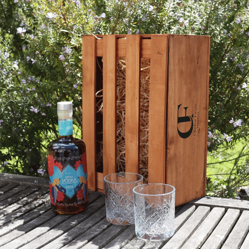 A bottle of Koakoa Coffee Vodka with a colorful label, alongside two clear pressed glass tumblers, all presented in a stained wooden gift box with a logo on the front.