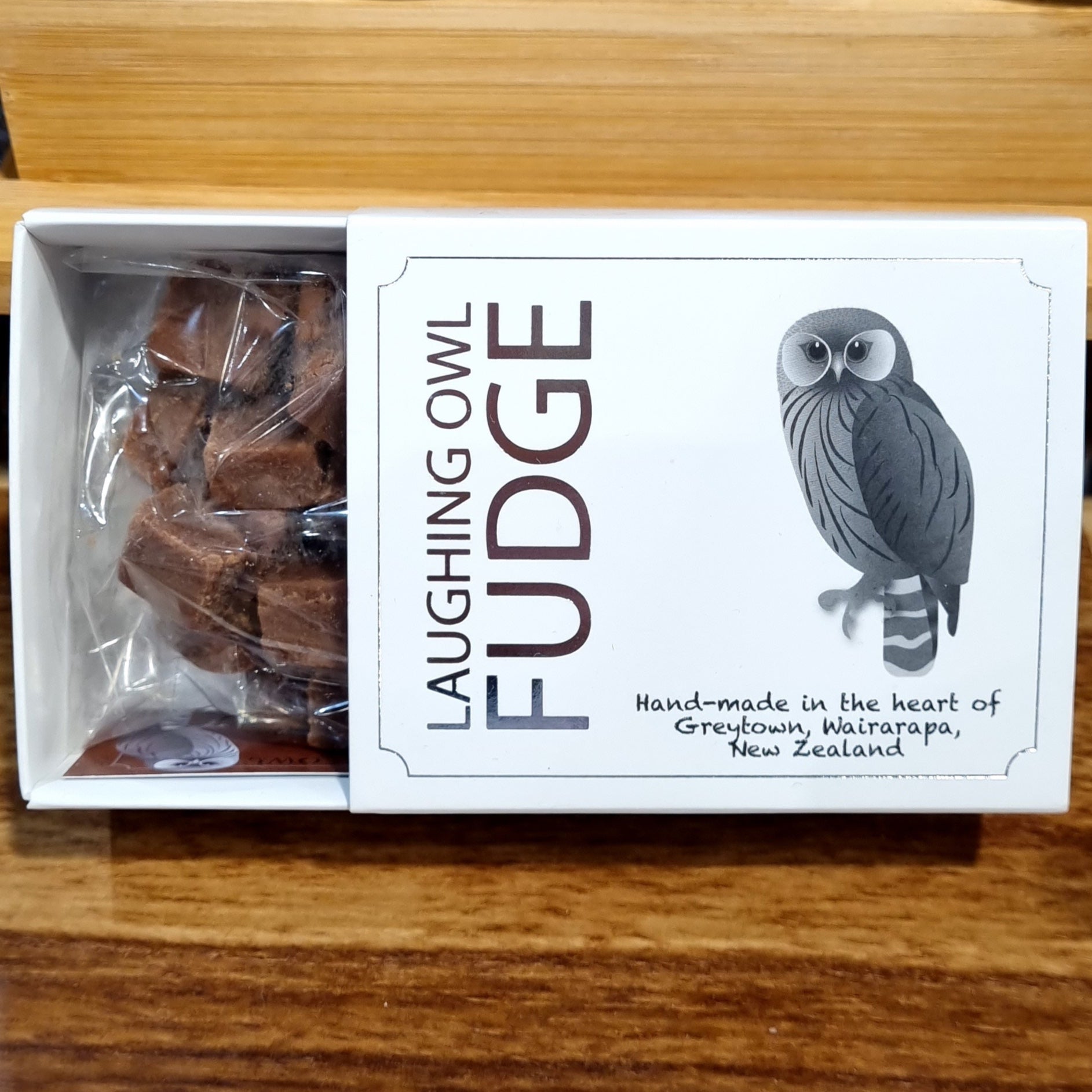 A gift box of Laughing Owl brand fudge, showing the box open to reveal individual pieces of fudge wrapped in transparent paper. The box has silver embossing with an owl illustration and text.