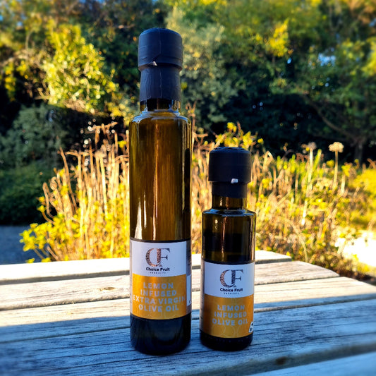 Two bottles of ChoiceFruits brand lemon infused olive oil, one larger and one smaller, placed on a table outdoors with foliage in the background.