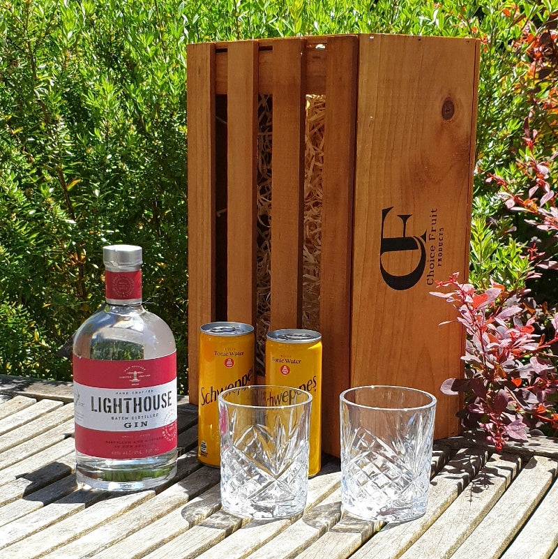A bottle of Lighthouse Gin with two tonic cans and two empty glass tumblers in front of a wooden gift box, all presented on a wooden surface outdoors.