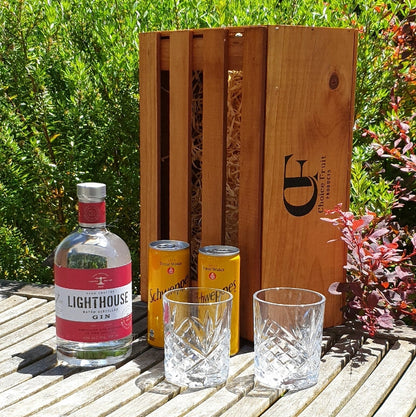 A bottle of Lighthouse Gin with two tonic cans and two empty glass tumblers in front of a wooden gift box, all presented on a wooden surface outdoors.