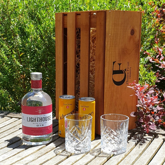 A bottle of Lighthouse Gin with two tonic cans and two empty glass tumblers in front of a wooden gift box, all presented on a wooden surface outdoors.