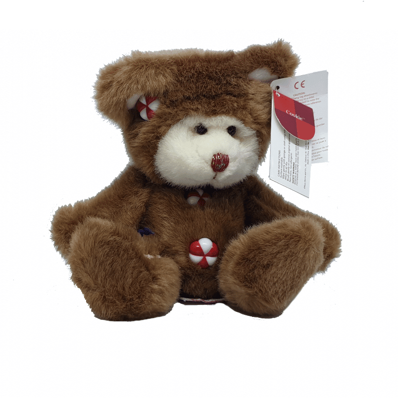 A plush teddy bear designed to look like a gingerbread man, with brown fur and white and red accents, sitting upright with a tag attached to its ear.