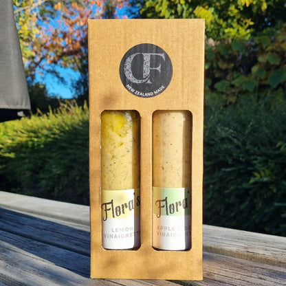 A gift box containing two bottles of vinaigrette sauce, one labeled 'Flora' and another with a visible lemon dressing, placed on a wooden surface with foliage in the background.