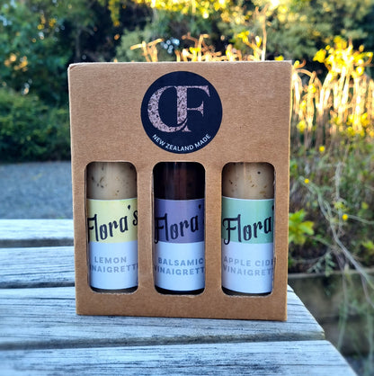A gift box containing three bottles of Flora's vinaigrettes in different flavors placed on a wooden surface with foliage in the background.